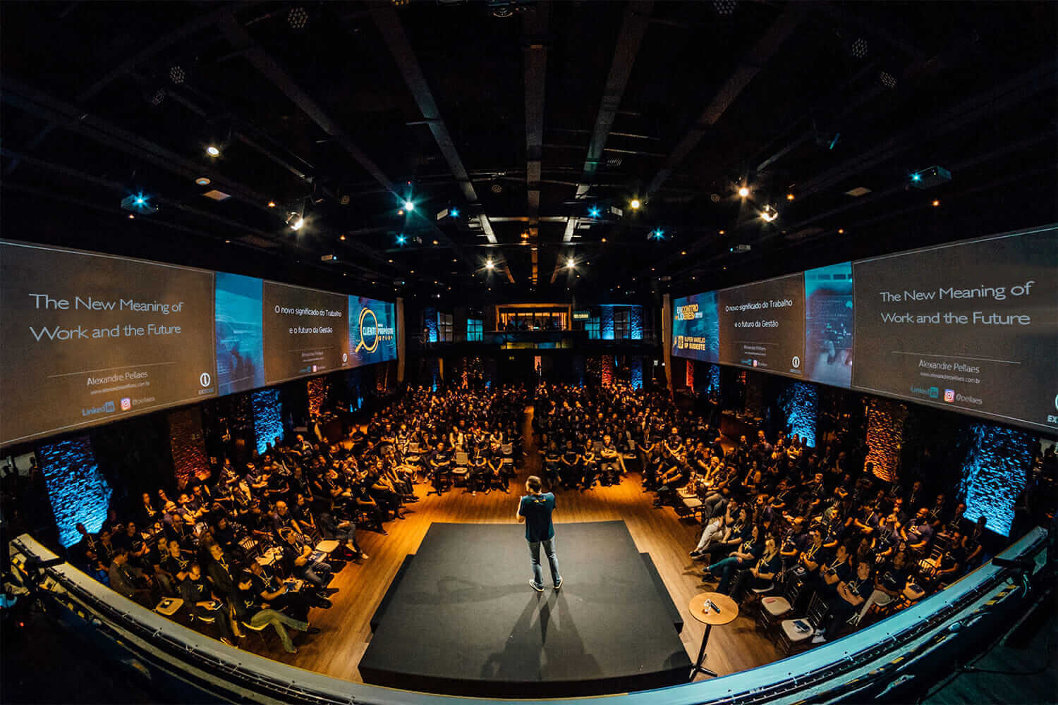 Fisheye view of a large conference with speaker on stage, audience seated, and presentation screens displaying "The New Meaning of Work."